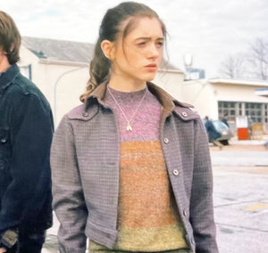 A young woman with long brown hair tied back, wearing the Zing Apparel Stranger Things S01 Nancy Wheeler Purple Jacket over a striped sweater and a necklace, stands outdoors in a parking lot with cars and buildings behind her.