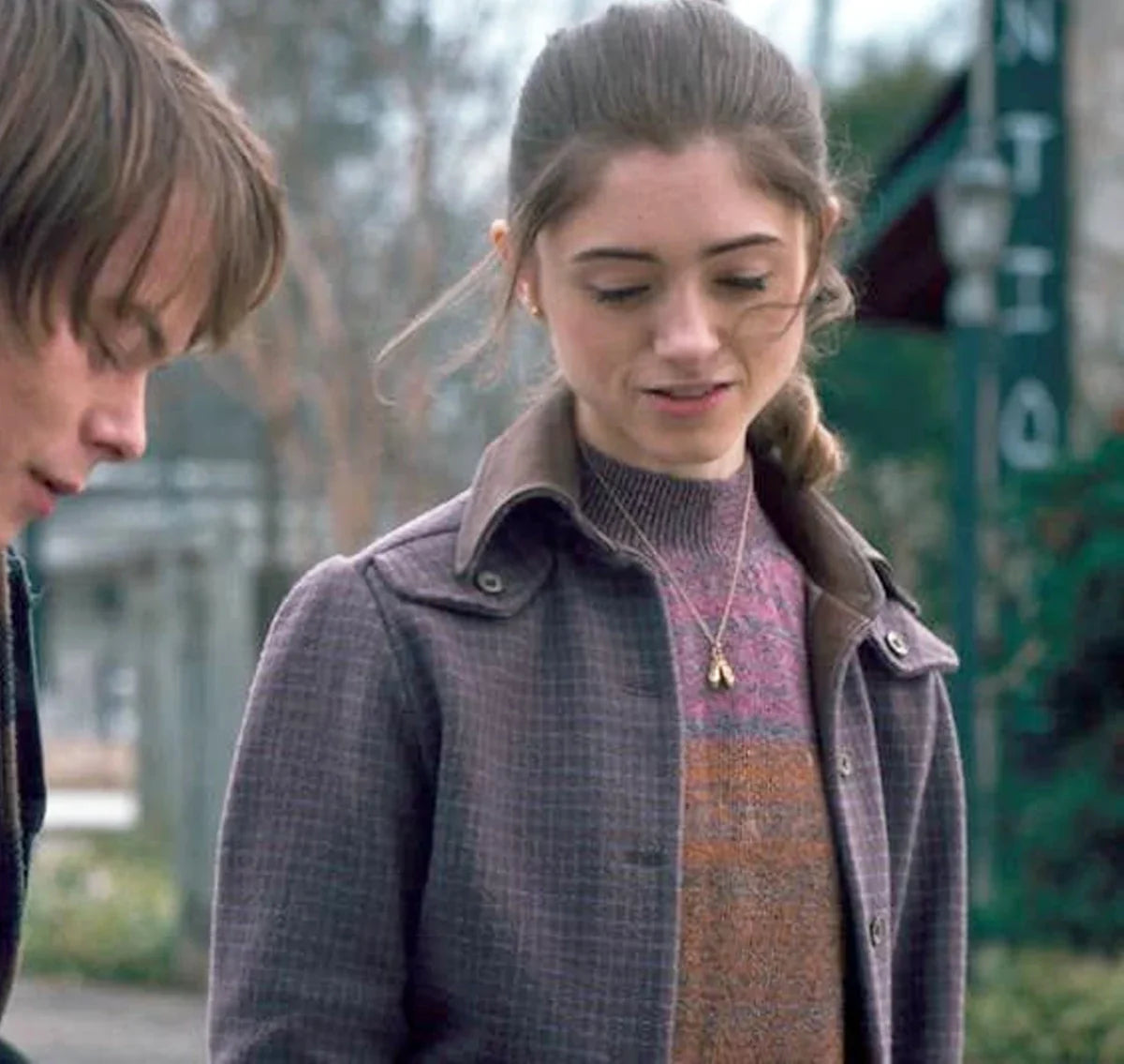 A young woman with light brown hair smiles while wearing the Stranger Things S01 Nancy Wheeler Purple Jacket by Zing Apparel, paired with a striped sweater and gold pendant necklace. Outdoors, a young man with brown hair is partly visible beside her.